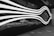 A futuristic and abstract design featuring white, wave-like lines on a dark reflective surface. The lines appear to create a fluid, motion-like effect, and a car can be seen reflected in the glossy surface beneath the lines. The setting appears to be outdoors, possibly near an entrance to a building.
