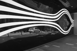 A futuristic and abstract design featuring white, wave-like lines on a dark reflective surface. The lines appear to create a fluid, motion-like effect, and a car can be seen reflected in the glossy surface beneath the lines. The setting appears to be outdoors, possibly near an entrance to a building.