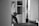 A person stands in what appears to be a gym or fitness room holding a medicine ball. The background features shelving with various items, including a plant. Exercise equipment like a TRX band is visible, as well as a water bottle and workout benches.