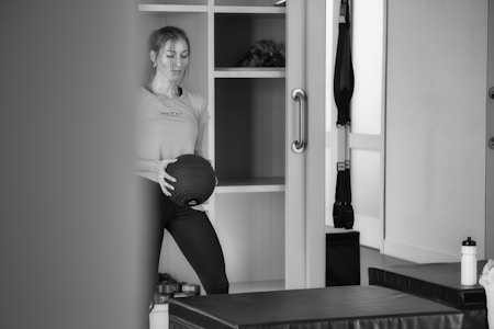 A person stands in what appears to be a gym or fitness room holding a medicine ball. The background features shelving with various items, including a plant. Exercise equipment like a TRX band is visible, as well as a water bottle and workout benches.