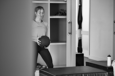 A person stands in what appears to be a gym or fitness room holding a medicine ball. The background features shelving with various items, including a plant. Exercise equipment like a TRX band is visible, as well as a water bottle and workout benches.