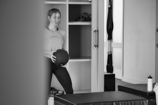 A person stands in what appears to be a gym or fitness room holding a medicine ball. The background features shelving with various items, including a plant. Exercise equipment like a TRX band is visible, as well as a water bottle and workout benches.