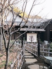 A traditional Japanese entrance gate marked with a white fabric displaying a symbol, likely related to a hot spring or onsen. The pathway is lined with wooden and bamboo fencing, leading to a rustic building with tiled roofing. Trees with bare branches frame the walkway, indicating a late autumn or winter setting. Fallen leaves and natural stones add to the serene and rustic ambiance.