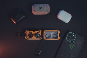 A collection of wireless earbuds in their cases and a smartphone in a protective case are arranged on a dark surface. The earbuds include various brands, and the smartphone features a distinctive logo on its case. The lighting is subdued, creating a sleek and modern aesthetic.