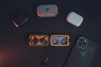 A collection of wireless earbuds in their cases and a smartphone in a protective case are arranged on a dark surface. The earbuds include various brands, and the smartphone features a distinctive logo on its case. The lighting is subdued, creating a sleek and modern aesthetic.