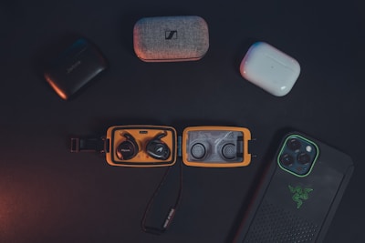 A collection of wireless earbuds in their cases and a smartphone in a protective case are arranged on a dark surface. The earbuds include various brands, and the smartphone features a distinctive logo on its case. The lighting is subdued, creating a sleek and modern aesthetic.