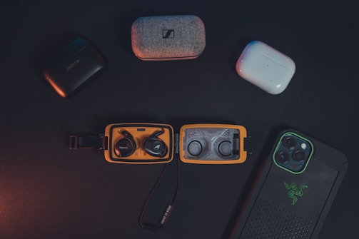 A collection of wireless earbuds in their cases and a smartphone in a protective case are arranged on a dark surface. The earbuds include various brands, and the smartphone features a distinctive logo on its case. The lighting is subdued, creating a sleek and modern aesthetic.