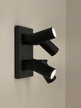 Two modern, black cylindrical spotlights are mounted on a wall. The design is sleek and minimalist, with the light beams directed downwards and slightly to the sides. The wall is a pale gray color, providing contrast to the dark fixtures.