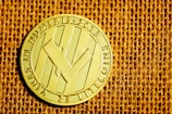 A shiny gold coin featuring a prominent logo or symbol in the center, with the text 'VIRES IN NUMERIS' and '2013' engraved along the edges. The coin rests on a textured burlap fabric background, with its intricate design and metallic sheen reflecting the light.