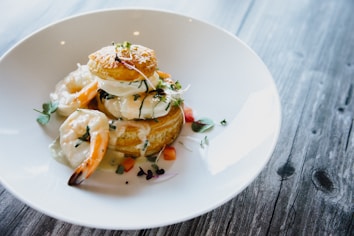 A white plate holds a gourmet dish featuring golden-brown pastry puffs topped with creamy sauce, surrounded by shrimp. Garnishes include small herb leaves and finely diced vegetables, adding color. The plate rests on a rustic wooden table.