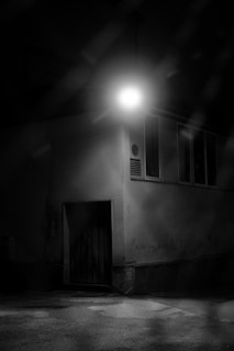 A dimly lit urban scene featuring a building corner under a solitary bright streetlight. The light casts stark shadows, creating a moody atmosphere. The weathered wall has some marks and textures, along with a closed wooden door at the bottom.