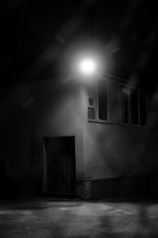 A dimly lit urban scene featuring a building corner under a solitary bright streetlight. The light casts stark shadows, creating a moody atmosphere. The weathered wall has some marks and textures, along with a closed wooden door at the bottom.