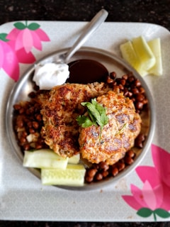 A metal plate holds two patties garnished with fresh cilantro. The dish is surrounded by cooked chickpeas and creamy sauces, with cucumber slices on the side. The plate is situated on a decorative tray with pink floral patterns.