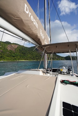 A luxurious yacht sailing near lush green hills under a partly cloudy sky. The yacht deck is spacious with cushioned sunbeds and a visible steering area. The calm, clear water contrasts beautifully with the greenery and blue sky.