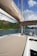 A luxurious yacht sailing near lush green hills under a partly cloudy sky. The yacht deck is spacious with cushioned sunbeds and a visible steering area. The calm, clear water contrasts beautifully with the greenery and blue sky.