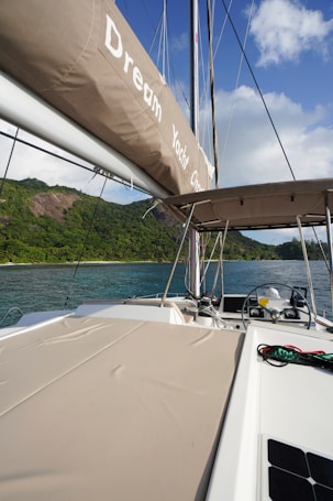 A luxurious yacht sailing near lush green hills under a partly cloudy sky. The yacht deck is spacious with cushioned sunbeds and a visible steering area. The calm, clear water contrasts beautifully with the greenery and blue sky.