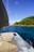 A sailboat deck featuring cushioned seating, a solar panel, and fishing rods. The boat is situated on calm blue waters with an island covered in lush green trees and rocky hills in the background under a clear blue sky.