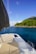 A sailboat deck featuring cushioned seating, a solar panel, and fishing rods. The boat is situated on calm blue waters with an island covered in lush green trees and rocky hills in the background under a clear blue sky.