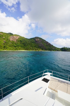 A beautiful, lush green island extends across the horizon, bordered by clear blue ocean waters. The foreground features the bow of a white yacht, with comfortable seating visible, providing a view of both the sea and the scenic landscape. Fluffy white clouds sporadically dot the blue sky.