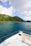 A beautiful, lush green island extends across the horizon, bordered by clear blue ocean waters. The foreground features the bow of a white yacht, with comfortable seating visible, providing a view of both the sea and the scenic landscape. Fluffy white clouds sporadically dot the blue sky.