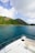 A beautiful, lush green island extends across the horizon, bordered by clear blue ocean waters. The foreground features the bow of a white yacht, with comfortable seating visible, providing a view of both the sea and the scenic landscape. Fluffy white clouds sporadically dot the blue sky.
