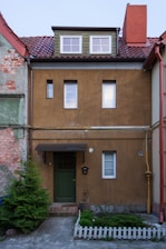 A two-story townhouse with a brown facade and a sloped red-tiled roof. The house features three windows on the upper floor and one window next to a green door on the ground floor. There is a small garden with plants surrounded by a white picket fence in front of the house. The walls show signs of aging with some exposed brick and peeling paint.
