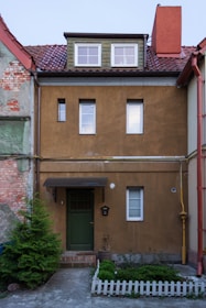 A two-story townhouse with a brown facade and a sloped red-tiled roof. The house features three windows on the upper floor and one window next to a green door on the ground floor. There is a small garden with plants surrounded by a white picket fence in front of the house. The walls show signs of aging with some exposed brick and peeling paint.