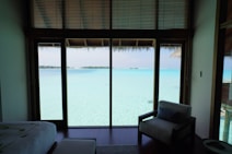 A serene view from inside a room featuring a window overlooking a turquoise blue ocean. The room contains a bed with white linens and an armchair. The sunlight casts gentle reflections from the clear water.