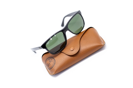 A pair of black-framed sunglasses with green-tinted lenses rests on a brown leather case. The case features a snap closure and embossed branding.