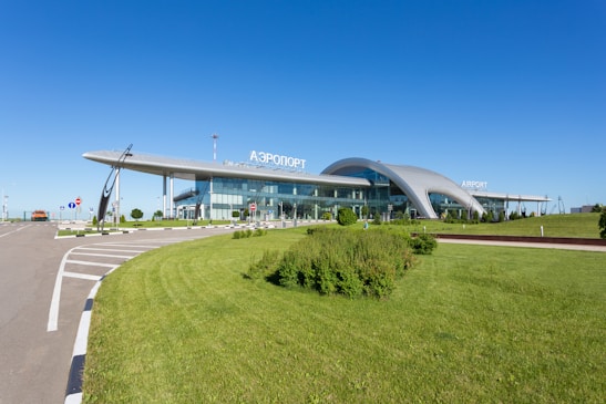 A modern airport terminal with a sleek, curving roof and extensive glass facade. There is a clear blue sky above, lush green grass in the foreground, and various signs along the roadway leading to the terminal.