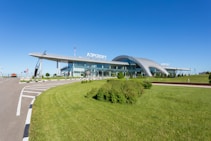 A modern airport terminal with a sleek, curving roof and extensive glass facade. There is a clear blue sky above, lush green grass in the foreground, and various signs along the roadway leading to the terminal.