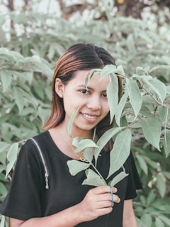 A woman is smiling and holding a branch with green leaves in front of leafy greenery. She has brown hair and is wearing a black top.
