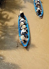 A group of people in traditional conical hats are paddling along a murky river in long, narrow blue boats. They are navigating through a waterway surrounded by lush, green foliage.