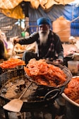 A person with a long beard, wearing a blue turban, is preparing a batch of fried sweets, likely jalebi, in a busy market setting. The sweets are orange and are being lifted from the hot oil with a large spatula, dripping with sugar syrup.