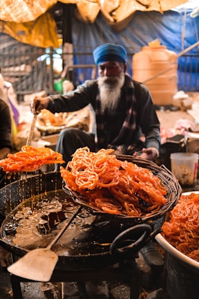 A person with a long beard, wearing a blue turban, is preparing a batch of fried sweets, likely jalebi, in a busy market setting. The sweets are orange and are being lifted from the hot oil with a large spatula, dripping with sugar syrup.