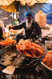 A person with a long beard, wearing a blue turban, is preparing a batch of fried sweets, likely jalebi, in a busy market setting. The sweets are orange and are being lifted from the hot oil with a large spatula, dripping with sugar syrup.