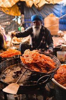 A person with a long beard, wearing a blue turban, is preparing a batch of fried sweets, likely jalebi, in a busy market setting. The sweets are orange and are being lifted from the hot oil with a large spatula, dripping with sugar syrup.