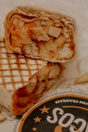 A close-up of a grilled wrap filled with pieces of chicken and melted cheese, with grill marks visible on the tortilla. The wrap is placed on a piece of branded paper.