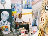 An artist sits in a studio surrounded by various colorful paintings and art supplies. The room is filled with vibrant canvases depicting portraits and abstract designs, creating a lively and creative atmosphere.