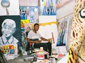 An artist sits in a studio surrounded by various colorful paintings and art supplies. The room is filled with vibrant canvases depicting portraits and abstract designs, creating a lively and creative atmosphere.