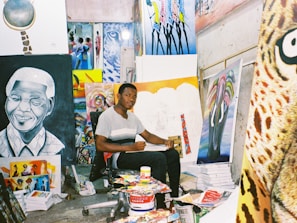 An artist sits in a studio surrounded by various colorful paintings and art supplies. The room is filled with vibrant canvases depicting portraits and abstract designs, creating a lively and creative atmosphere.