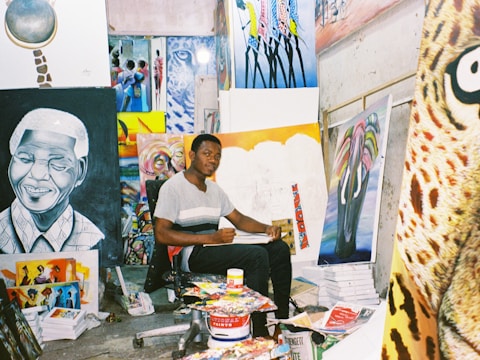 An artist sits in a studio surrounded by various colorful paintings and art supplies. The room is filled with vibrant canvases depicting portraits and abstract designs, creating a lively and creative atmosphere.