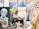 An artist sits in a studio surrounded by various colorful paintings and art supplies. The room is filled with vibrant canvases depicting portraits and abstract designs, creating a lively and creative atmosphere.