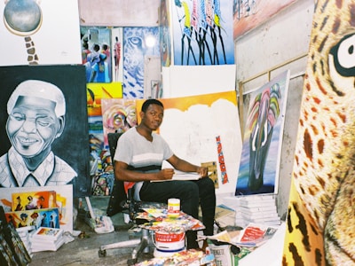 An artist sits in a studio surrounded by various colorful paintings and art supplies. The room is filled with vibrant canvases depicting portraits and abstract designs, creating a lively and creative atmosphere.