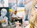 An artist sits in a studio surrounded by various colorful paintings and art supplies. The room is filled with vibrant canvases depicting portraits and abstract designs, creating a lively and creative atmosphere.