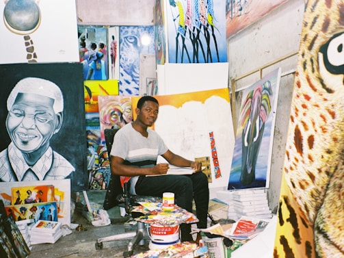 An artist sits in a studio surrounded by various colorful paintings and art supplies. The room is filled with vibrant canvases depicting portraits and abstract designs, creating a lively and creative atmosphere.