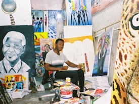 An artist sits in a studio surrounded by various colorful paintings and art supplies. The room is filled with vibrant canvases depicting portraits and abstract designs, creating a lively and creative atmosphere.