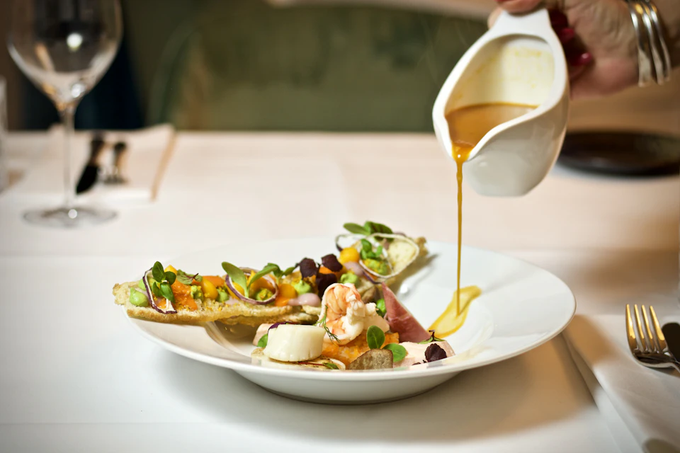 Woman holds pitcher with sauce over seafood dish at a dining table setting