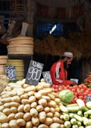 A market scene featuring various vegetables displayed prominently, including a large pile of potatoes in the foreground and other produce like tomatoes, a cabbage, and cucumbers. A vendor wearing a red jacket and cap is seen on the right side, possibly noting something down. The background is filled with baskets and other market items, creating a rustic and bustling atmosphere.