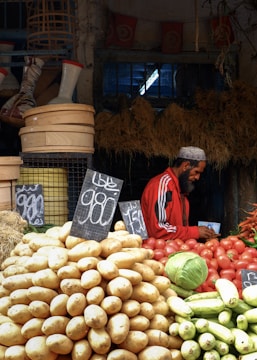 A market scene featuring various vegetables displayed prominently, including a large pile of potatoes in the foreground and other produce like tomatoes, a cabbage, and cucumbers. A vendor wearing a red jacket and cap is seen on the right side, possibly noting something down. The background is filled with baskets and other market items, creating a rustic and bustling atmosphere.